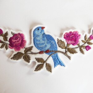 Puede incluir: Parche bordado con un pájaro azul posado en una rama con rosas y capullos rosas. El parche tiene un fondo blanco y costuras detalladas. El pájaro tiene pico y patas rojas.