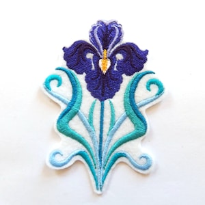 May include: Embroidered patch featuring a stylized iris flower in shades of deep blue and teal. The flower has a detailed design with yellow accents, set against a white background. The patch has a decorative, Art Nouveau-inspired design.