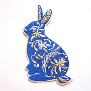 May include: Blue embroidered rabbit patch with white and gold floral details. The rabbit is sitting with its ears up and its tail behind it.