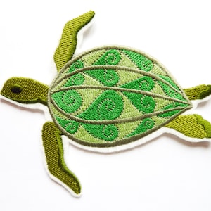 May include: An embroidered patch in the shape of a sea turtle. The turtle features a light green shell with a dark green swirling leaf-like pattern. The head and flippers are a solid olive green, and the entire patch has a white border.