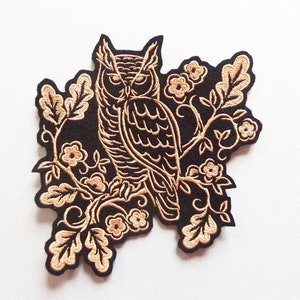 Könnte beinhalten: Gestickter Bügel-Patch mit einer schwarzen Eule mit goldenen Nähten, die auf einem Zweig mit goldenen Blättern und Blüten sitzt.