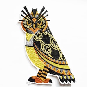 Peut inclure: Un patch brodé en forme de hibou, de couleur jaune, orange, noir et gris. Le hibou a un design détaillé avec des motifs géométriques et une expression féroce.