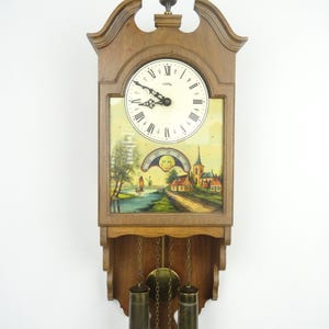 May include: A wooden wall clock featuring a cream-coloured face with black Roman numerals and ornate hands. Below the clock face is a painted landscape scene with a moon phase dial. Two brass weights hang from chains beneath the clock body.
