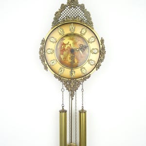 May include: A wall clock featuring an ornate gold-toned metal frame with intricate scrollwork and a central circular dial. The dial displays a painted scene of figures in a landscape, surrounded by gold hour markers and black hands, with two hanging brass weights.