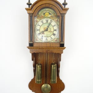 May include: A wooden wall clock with a detailed face featuring a sailboat scene, Roman numerals, and a sun and moon design. The clock has a pendulum and two brass weights, with decorative finials and a carved wooden frame.