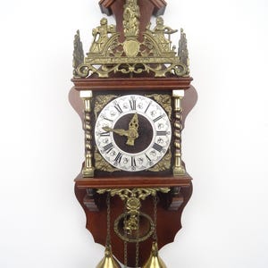 May include: A vintage wall clock with a dark wood frame and ornate brass accents. The clock face features Roman numerals and a decorative center. Two brass pendulum weights hang below. The clock is approximately 24 inches tall.