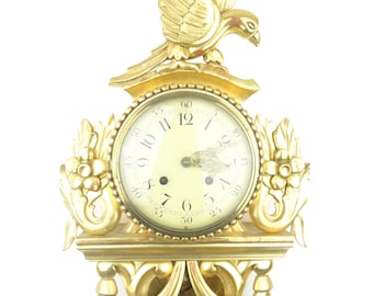 May include: A gold-colored wall clock with a bird perched on top. The clock face has black numerals and hands. Ornate floral and scrollwork details frame the clock face and base. The clock is approximately 12 inches tall.