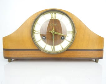 May include: A vintage wooden mantel clock with a light brown finish. The clock face has Roman numerals and gold-coloured hands. The clock is marked "Hermle" and has a curved top.