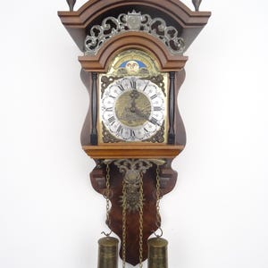 May include: A wooden wall clock featuring a brass dial with Roman numerals, a moon phase indicator, and an engraved landscape scene. The clock has two hanging brass weights suspended by chains, decorative metal scrollwork, and three finials on top.