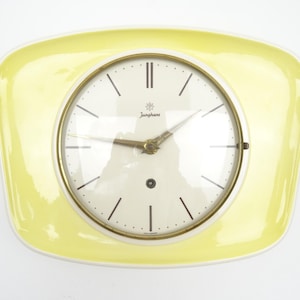 Puede incluir: Un reloj de pared con un marco amarillo pálido de forma rectangular redondeada y una esfera circular blanca. El reloj presenta marcadores de hora negros finos, manecillas doradas y un bisel dorado. El nombre Junghans está impreso en negro.