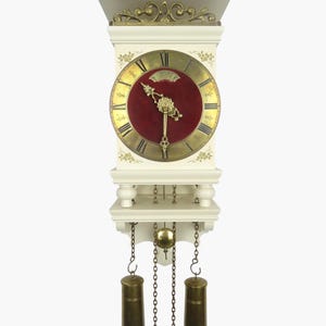 Könnte beinhalten: Eine cremefarbene Wanduhr mit rotem Zifferblatt und goldenen Zeigern und Ziffern. Die Uhr hat dekorative Details an der Ober- und Unterseite, mit zwei zylindrischen Gewichten, die darunter hängen. Abmessungen sind nicht sichtbar.