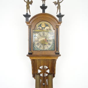 May include: A wooden wall clock with a decorative design. The clock features a round face with Roman numerals, a painted landscape scene, and three bronze figures on top. The clock has a pendulum and weights.