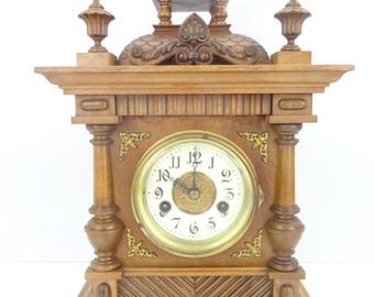 May include: A wooden mantel clock with a light brown finish, featuring a circular white face with black Arabic numerals and ornate gold-tone corner accents. The clock has a decorative top structure with finials and a central dome element.