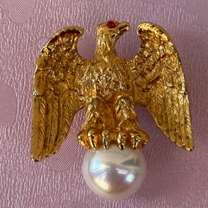 May include: A gold-toned eagle brooch with outstretched wings, a red gemstone eye, and a lustrous pearl. The eagle is perched atop the pearl, creating a striking contrast. The brooch is likely made of metal and is a decorative accessory.