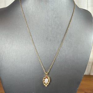 May include: A gold-toned necklace with a pendant featuring a central oval-shaped, off-white stone. The pendant is framed by small red and yellow-toned accents. The necklace is displayed on a black mannequin.