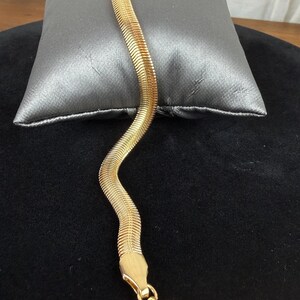 May include: A gold-colored herringbone bracelet with a lobster clasp. The bracelet is displayed on a gray pillow against a black background. The bracelet has a flat, snake-like design.