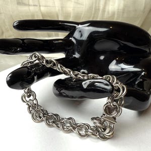 May include: A silver chain bracelet with a double-link design. The bracelet is on a black hand-shaped display stand.