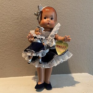 May include: A porcelain doll wearing a white and blue dress with a lace trim. The doll is holding a smaller porcelain doll wearing a blue dress. The larger doll has a tag that says "I am Patsy".