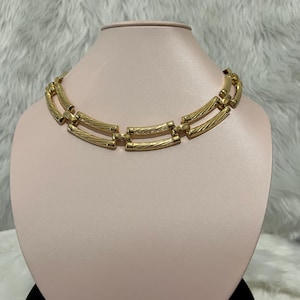 May include: A gold-toned necklace with a double row of textured, rectangular links. The necklace is displayed on a pink mannequin bust.
