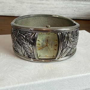 May include: A silver-toned cuff watch featuring a textured leaf pattern on the band and a rectangular face with a light-colored dial. The watch face displays the text Peek & Peek and the word Quartz, with gold-colored hands and hour markers.