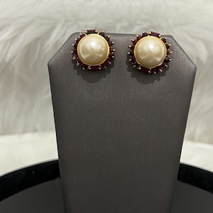 May include: A pair of gold tone earrings with a large faux pearl center and a circle of red rhinestones around the pearl.