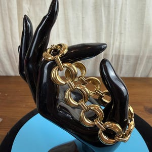 May include: A gold-toned bracelet with large, interlocking circular links. The bracelet is displayed on a black hand-shaped jewelry holder against a light blue and black background. The bracelet's design features a secure clasp.