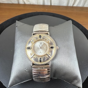 Op de afbeelding: Zilverkleurig polshorloge met een ronde wijzerplaat met Romeinse cijfers en een decoratieve rand. Het horloge heeft een zilveren, rekbare band en zwarte accenten. Het horloge wordt gepresenteerd op een grijs kussen.