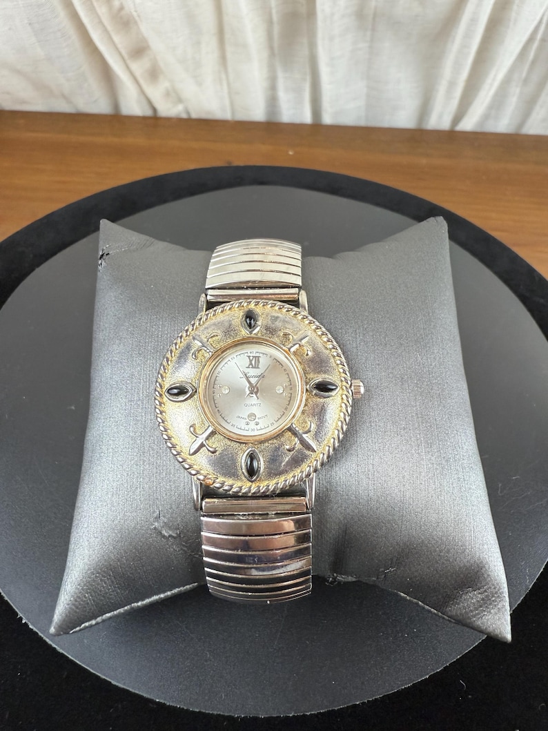 Op de afbeelding: Zilverkleurig polshorloge met een ronde wijzerplaat met Romeinse cijfers en een decoratieve rand. Het horloge heeft een zilveren, rekbare band en zwarte accenten. Het horloge wordt gepresenteerd op een grijs kussen.