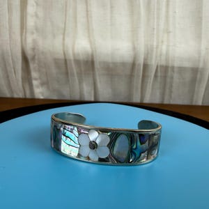 May include: Silver cuff bracelet with iridescent abalone shell inlay and a white flower design. The bracelet has a rectangular shape with rounded edges. The flower has a dark center. The bracelet is on a blue surface.