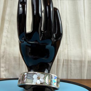 May include: A silver cuff bracelet with an abalone and mother-of-pearl inlay. The bracelet features a white flower design with a central gemstone. The bracelet is displayed on a black hand-shaped jewelry holder.