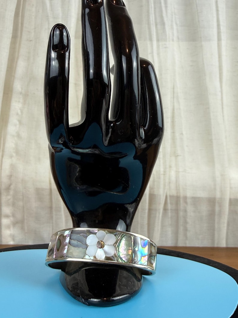 May include: A silver cuff bracelet with an abalone and mother-of-pearl inlay. The bracelet features a white flower design with a central gemstone. The bracelet is displayed on a black hand-shaped jewelry holder.