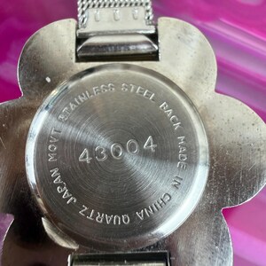 Może przedstawiać: Zbliżenie srebrnego zegarka kwarcowego z tarczą w kształcie kwiatu. Zegarek ma stalowy tył z numerem 43004 i napisem "Made in China". Pasek zegarka jest srebrny.