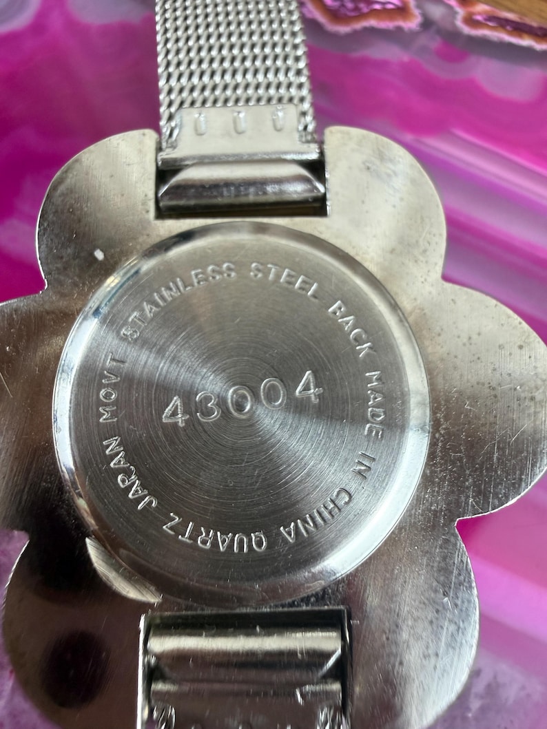 Może przedstawiać: Zbliżenie srebrnego zegarka kwarcowego z tarczą w kształcie kwiatu. Zegarek ma stalowy tył z numerem 43004 i napisem "Made in China". Pasek zegarka jest srebrny.