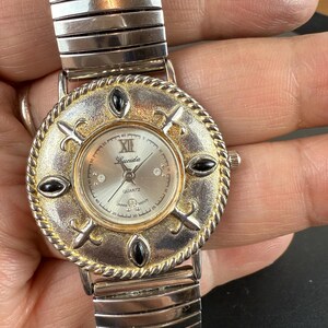 Op de afbeelding: Lucida Quartz horloge met een zilveren wijzerplaat, goudkleurige lunette en zwarte accenten. Het horloge heeft een zilverkleurige, rekbare band. De wijzerplaat heeft Romeinse cijfers en is versierd met decoratieve elementen. Vintage stijl.