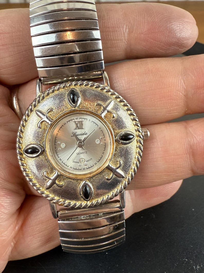 Op de afbeelding: Lucida Quartz horloge met een zilveren wijzerplaat, goudkleurige lunette en zwarte accenten. Het horloge heeft een zilverkleurige, rekbare band. De wijzerplaat heeft Romeinse cijfers en is versierd met decoratieve elementen. Vintage stijl.