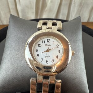 May include: A silver-toned quartz watch with a white face and black numerals. The watch has a round face with the brand name "Ronica" printed on the face. The watch is displayed on a gray pillow.