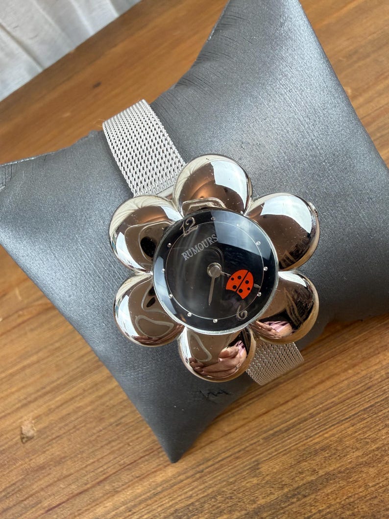 Może przedstawiać: Srebrny zegarek z tarczą w kształcie kwiatu i siatkowym paskiem. Czarna tarcza zegarka ma czerwony wz&oacute;r biedronki i napis "RUMOURS". Zegarek jest prezentowany na szarej poduszce.