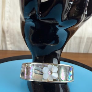 May include: A silver-toned cuff bracelet with iridescent abalone shell inlays. The bracelet features a white flower with a central gemstone. The bracelet is displayed on a black hand-shaped stand.