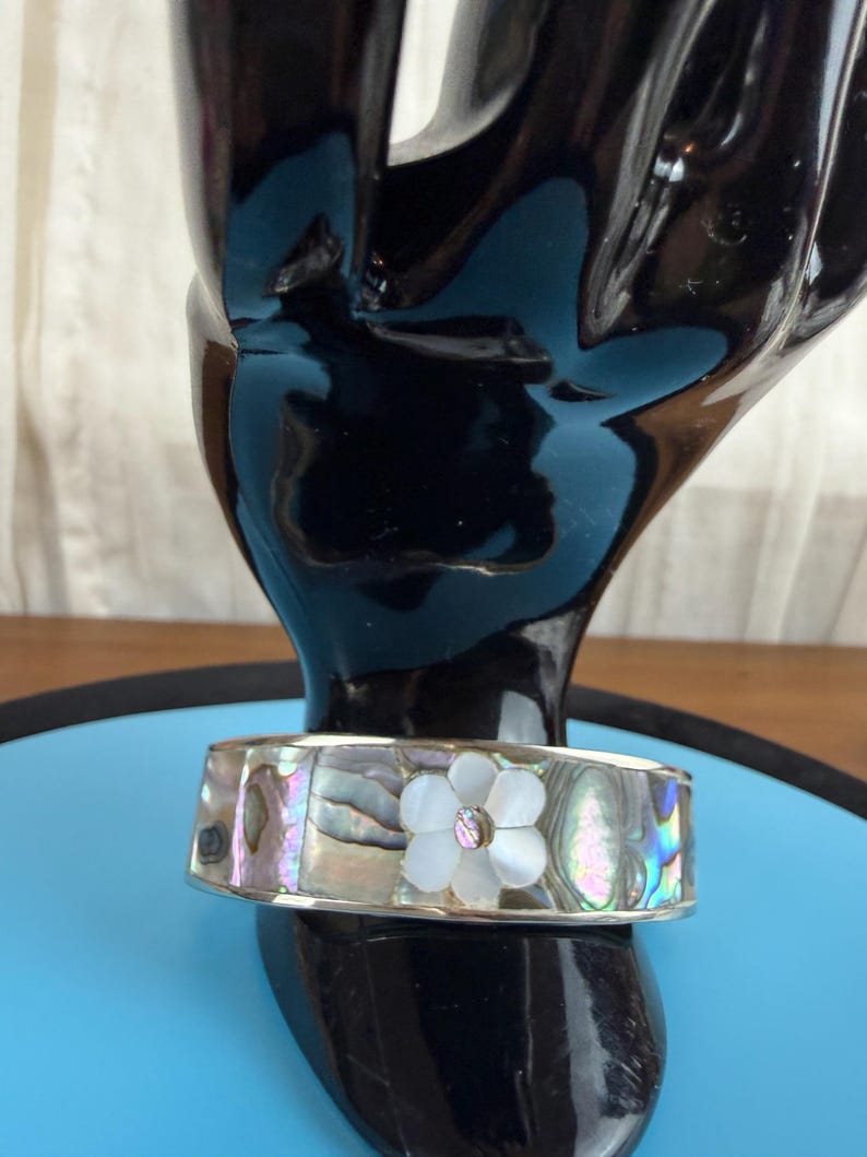 May include: A silver-toned cuff bracelet with iridescent abalone shell inlays. The bracelet features a white flower with a central gemstone. The bracelet is displayed on a black hand-shaped stand.