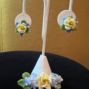 Op de afbeelding: Een paar porseleinen bloemoorbellen met gele rozen en blauwe vergeet-me-nietjes. De oorbellen worden gepresenteerd op een witte standaard.