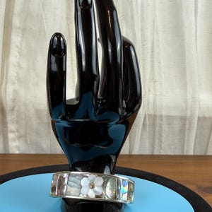 May include: A silver bracelet with a floral design. The bracelet features a mother-of-pearl flower with a central gem. The bracelet is displayed on a black hand-shaped stand against a blue background.