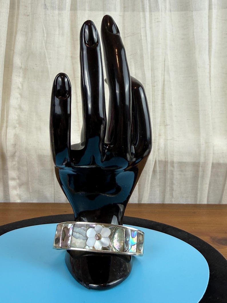 May include: A silver bracelet with a floral design. The bracelet features a mother-of-pearl flower with a central gem. The bracelet is displayed on a black hand-shaped stand against a blue background.