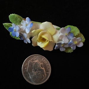 Op de afbeelding: Een porseleinen broche met een gele roos met blauwe en paarse vergeet-mij-nietjes. De broche is ongeveer zo groot als een Nederlandse tien-cent munt.