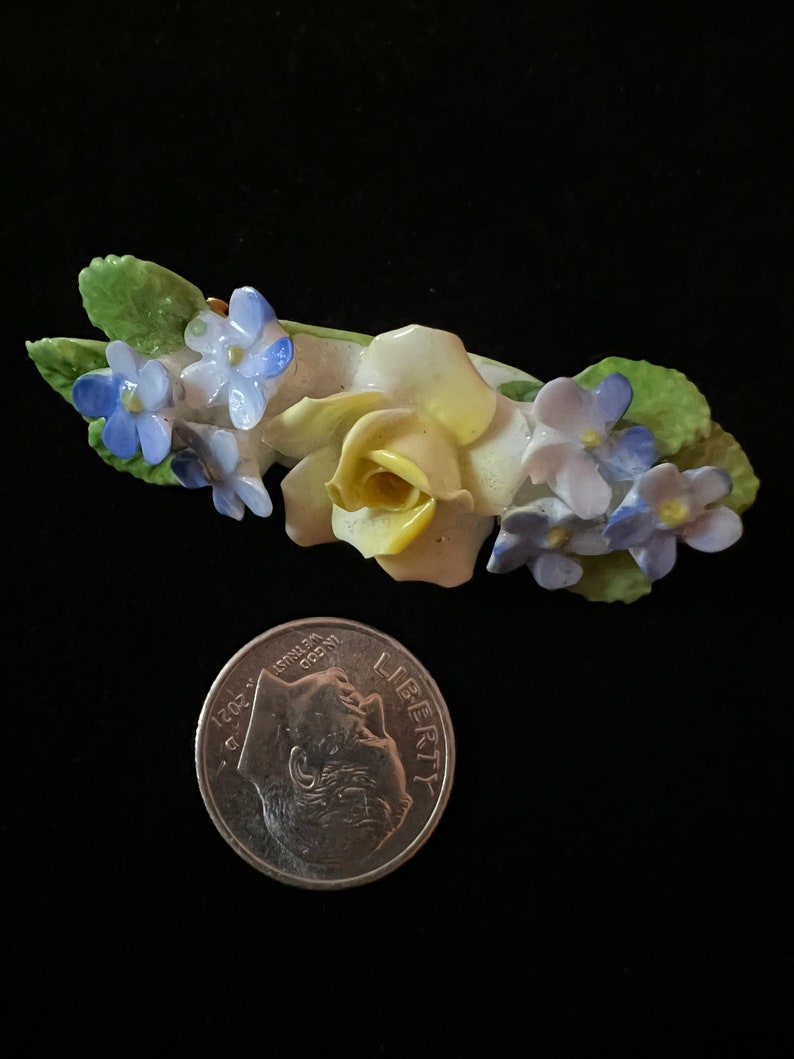Op de afbeelding: Een porseleinen broche met een gele roos met blauwe en paarse vergeet-mij-nietjes. De broche is ongeveer zo groot als een Nederlandse tien-cent munt.