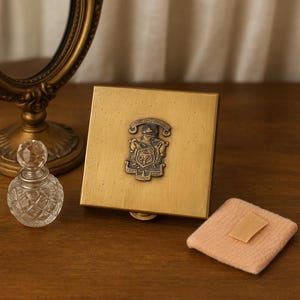 May include: A vintage gold-toned compact with a decorative emblem, a small glass perfume bottle, and a powder puff. The compact is square and has a hinged opening. The perfume bottle has a crystal stopper.