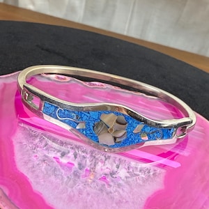 May include: Silver-tone bangle bracelet with a central decorative panel. The panel features a mosaic of blue and white stones, with a flower design in the center. The bracelet is set against a pink and white agate slice.