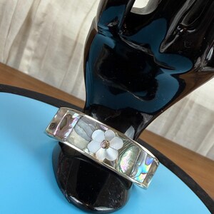 May include: A silver-tone bracelet with iridescent abalone shell inlays. The bracelet features a white flower with a small, clear center stone. The bracelet is displayed on a black hand-shaped stand against a blue background.