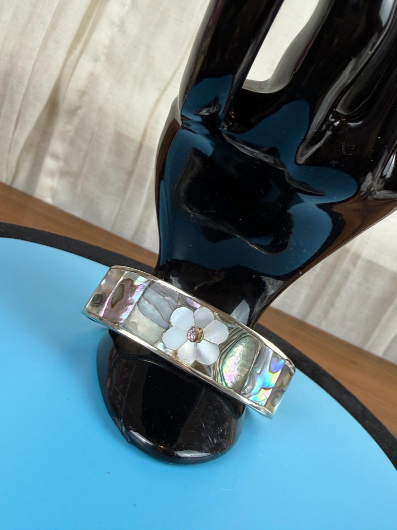 May include: A silver-tone bracelet with iridescent abalone shell inlays. The bracelet features a white flower with a small, clear center stone. The bracelet is displayed on a black hand-shaped stand against a blue background.