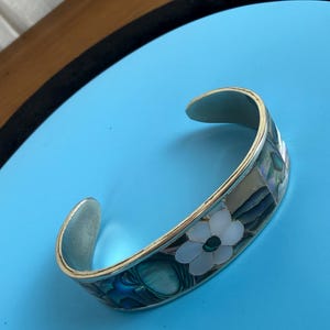 May include: Silver-toned cuff bracelet with a floral design. The bracelet features a white flower with a dark center, surrounded by iridescent blue and green accents. The bracelet is set against a blue background.
