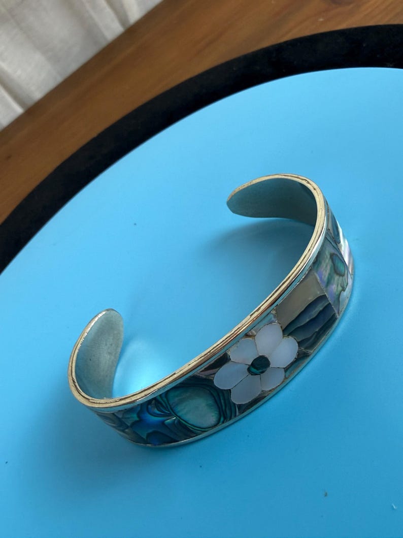 May include: Silver-toned cuff bracelet with a floral design. The bracelet features a white flower with a dark center, surrounded by iridescent blue and green accents. The bracelet is set against a blue background.
